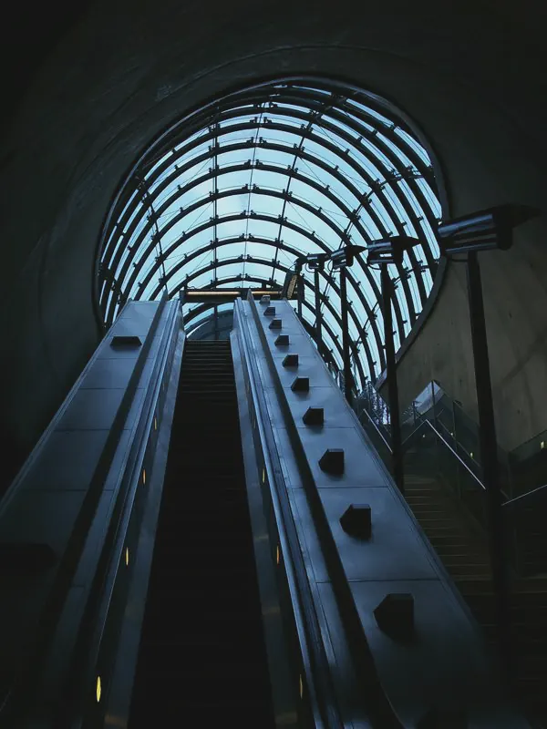 An escalator rises through a dark concrete tunnel towards a curved glass canopy of steel ribs and pale sky.