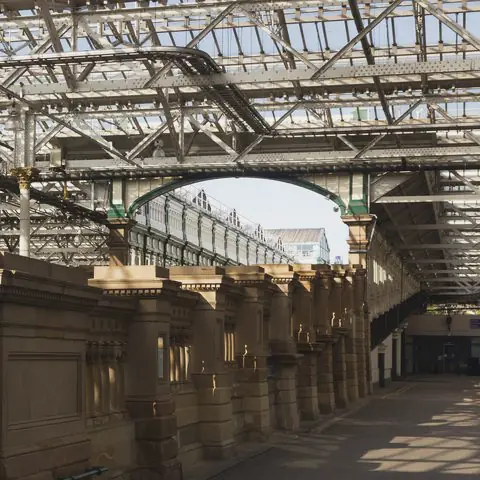 A steel and glass roof covers a stone colonnade that runs towards a bright arched opening.