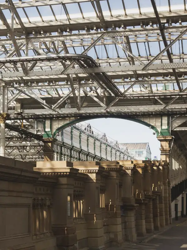 A steel and glass roof covers a stone colonnade that runs towards a bright arched opening.