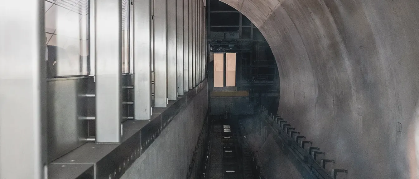 An inclined shaft drops away between steel ribs and a curved concrete wall towards a small lit window.