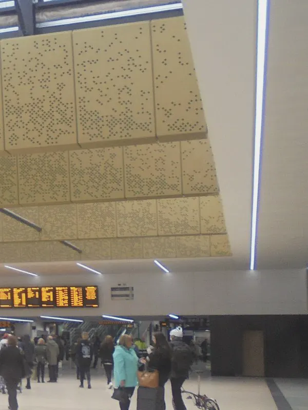 A gold perforated ceiling and skylights hang over the station concourse, with travellers small below.