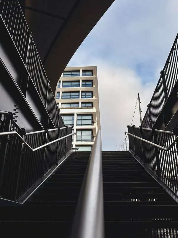 Black steel steps and a central handrail climb between railings towards a pale tower block and grey sky.