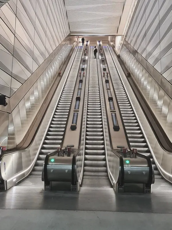 Three escalators climb a tall pale shaft lined with angled panels, a few riders small near the top.