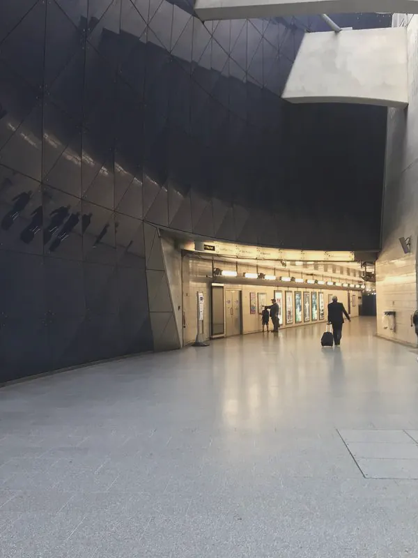 A curved blue glass wall sweeps towards a lit passage where a few travellers walk with luggage.