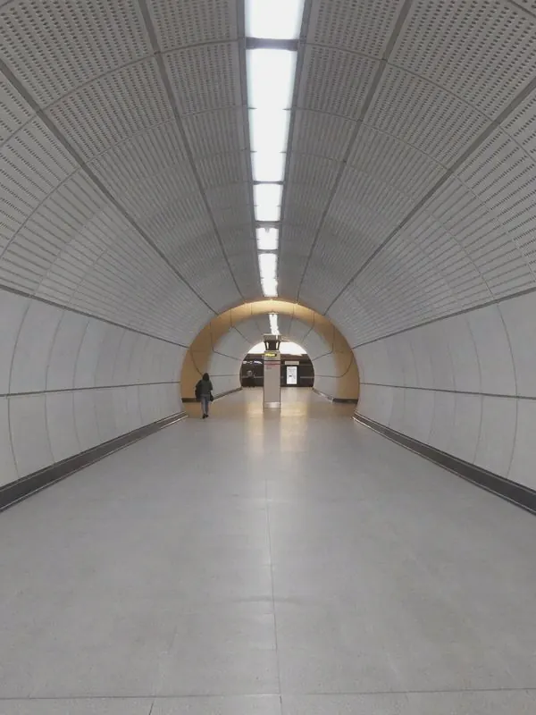 A long white tunnel of perforated panels runs straight ahead to a warm-lit arch and one distant walker.