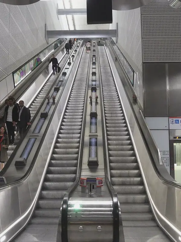 Three escalators climb a pale shaft between steel balustrades, a few riders small on the far left.