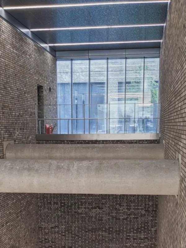 Escalators descend between tall brick walls beneath a concrete beam, with a glazed opening bright above.
